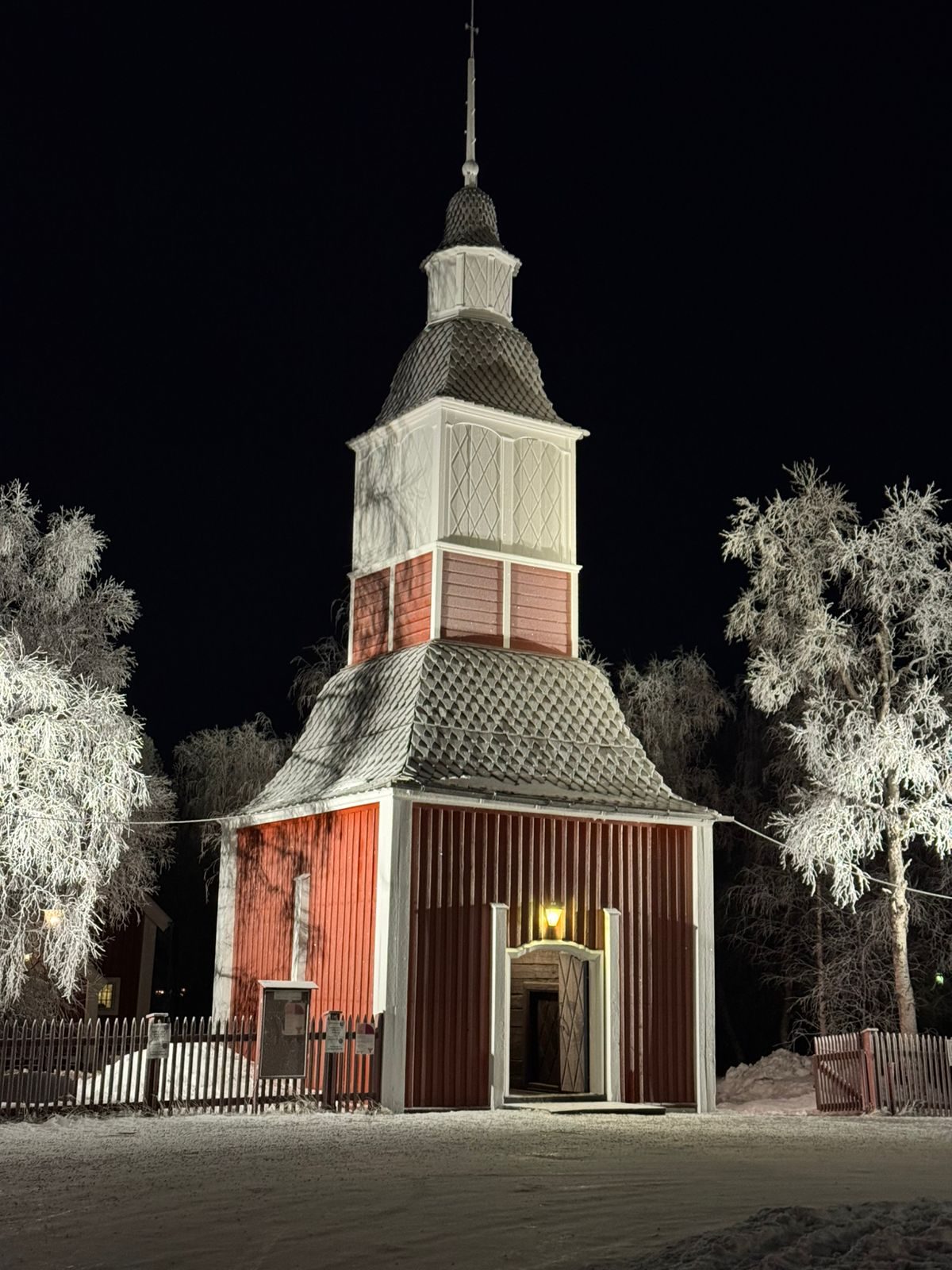 Campanário de madeira vermelha da igreja de Jukkasjärvi, iluminado à noite entre árvores cobertas de gelo.