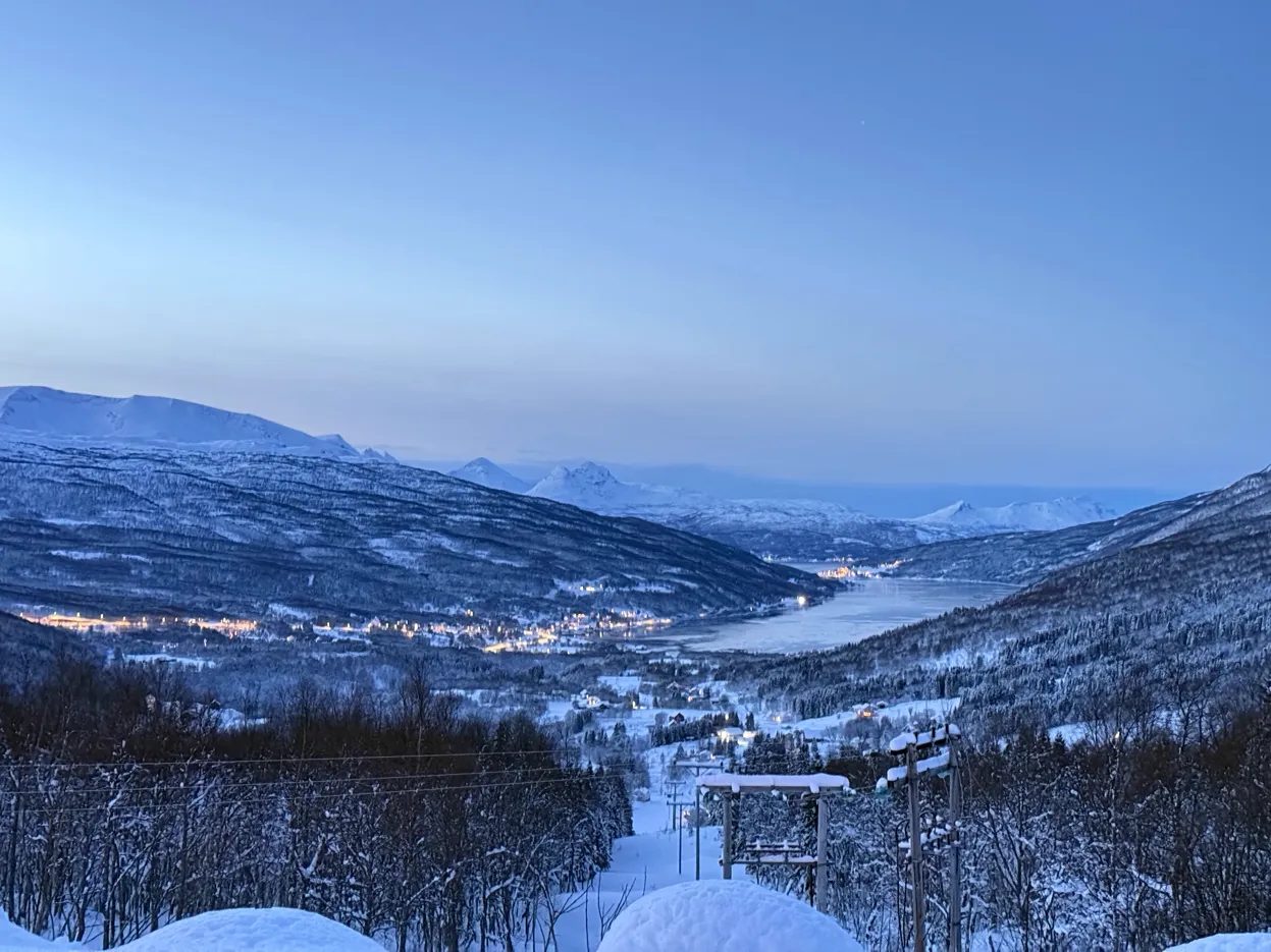 Vale coberto de neve ao entardecer azul, com as luzes de uma pequena cidade às margens da água.